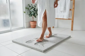Femme en robe de bain posant le pied sur un tapis de bain en diatomite gris dans une salle de bain familiale carrelée de blanc